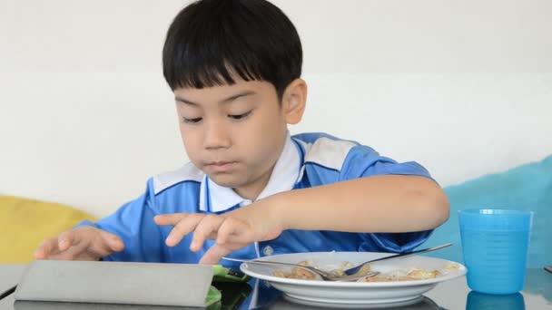 Little asian boy eating fried rice and playing tablet computer . — Stock Video