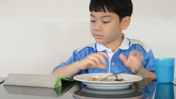 アジアの小さな男の子がチャーハンを食べ、タブレットコンピュータを遊ぶ . — ストック動画