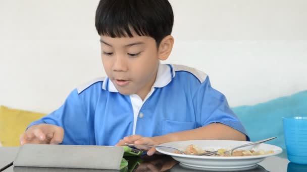 Little asian boy eating fried rice and playing tablet computer . — Stock Video