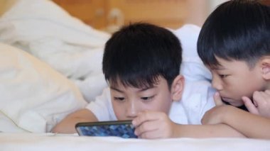 Asyalı çocuk gülümseyerek, yatakta döşeme tablet üzerinde oynuyor.