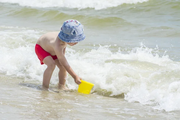 Niño recogiendo agua de mar en un cubo para el juego contra el telón de fondo de las olas — Foto de Stock