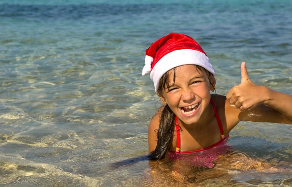 Niña divertida sonríe en una gorra de Santa Claus en la orilla del mar — Foto de Stock