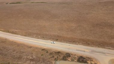 Büyüleyici hava görüntülerinde, ıssız bir yoldan geçen çarpıcı beyaz bir araba geniş ve kuru bir arazide zikzak çiziyor, macera ve keşif hissi uyandırıyor.