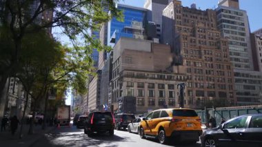 New York 10.28.2025. Güneşli şehir caddesi, yeşil ağaçlar, yoğun trafik ve New York 'ta valizlerle yürüyen bir yaya, seyahat ve şehir yaşam temaları için uygun parlak şehir atmosferi