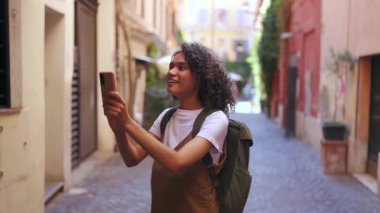 Sırt çantalı bir kadın akıllı telefon fotoğrafı çekerken dar renkli sokakları keşfediyor. Sıcak gün ışığının, rahat seyahat havasının ve şehir merkezinde yürüyüşü sırasında yumuşak sokak dokularının tadını çıkarıyor.