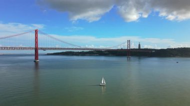 Lizbon Portekiz 'in hava görüntüsü 25 Nisan köprüsü, Tagus Nehri' ni kapsayan 25 de Abril Köprüsü, sakin su, tek yelkenli, uzak kıyı şeridi ve bulutlu mavi gökyüzü..