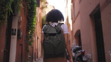 Eski binalarla çevrili dar bir şehir caddesinde yürürken sırt çantası takan bir kadının arka planı. Elinde telefon, sakin bir yolculuk ve şehir hayatı sahnesi yaratıyor..