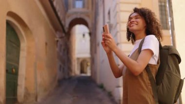Sırt çantasıyla gülümseyen kadın gezgin akıllı telefonunu tutuyor ve dar bir şehir caddesinde yürürken fotoğraf çekiyor rahat bir şehir gezisi sırasında anları yakalıyor..