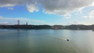 Lizbon Köprüsü 'nün Tagus nehri üzerindeki hava görüntüsü teknenin suları görünür bir şekilde kestiğini bulutlu gökyüzü süspansiyon kuleleri ve kıyılarda yumuşak güneş ışığı, Lizbon Portekiz