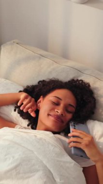 Sakin bir sabah sahnesinde, genç siyahi bir kadın yatakta yatarken akıllı telefonlara, gün ışığına ve beyaz yataklara göz gezdirirken rahat bir yaşam tarzı, yatakta dijital iletişim ve dinlenme konsepti yaratıyor.