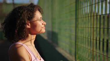 Tenis kortunun yanında uzanan pembe spor giysili atletik bir kadın, antrenmandan sonra dinlenirken yüzünde güneş ışığı, sakin ruh hali, açık hava fitness yaşam tarzı ve iyileşme anı..