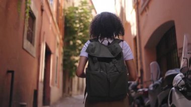 Sırt çantası takan genç bir kadın gezgin dar tarihi bir caddede yürüyor, çevredeki binalara bakarak kentsel çevreyi keşfediyor..