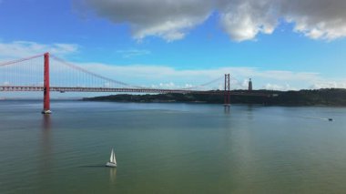 Lizbon Portekiz 'in 25 de Abril Köprüsü ile Tagus Nehri' ni kaplayan hava manzarası, sakin su yüzeyi, tek yelkenli, uzak nehir kıyıları ve dağınık bulutlu mavi gökyüzü..