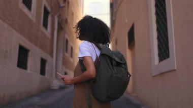 Sırt çantası taşıyan bir kadın gezgin telefonunu tutarken dar bir şehir caddesinde yürüyor, şehir çevresini keşfediyor ve yolculuğun tadını çıkarıyor..