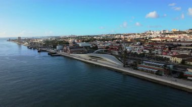 Drone kamera modern rıhtım binalarını, uzun nehir kenarındaki gezinti alanlarını, park edilmiş araçları ve berrak mavi gökyüzünün altında açık deniz kenarı ölçeğinde yumuşak yansımaları olan su yüzeyini gösteriyor.