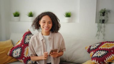 Modern oturma odasındaki kanepede otururken gülümseyen bir kadın dekoratif yastıklarla, gün ışığıyla, evde rahat rahat, rahat bir yaşam tarzıyla rahat bir şekilde oturuyor..