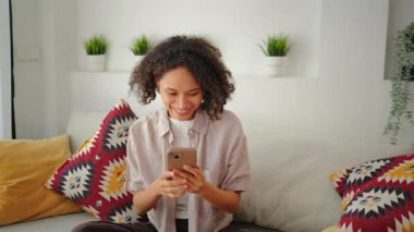 Kanepede bağdaş kurup oturan gülümseyen bir kadın akıllı telefon, rahat yastıklı parlak modern oturma odası, rahat gün ışığı, rahat yaşam tarzı anı ve her gün kapalı mekan sahnesi kullanıyor..