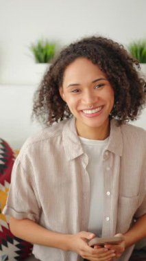 Kanepede oturan ve gülümseyen genç bir kadın kameraya ve yukarı bakarken, akıllı telefonu elinde tutarken, rahat ev içi, yumuşak doğal ışık, sakin yaşam tarzı sahnesi ve otantik günlük duygu..