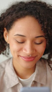 Kapalı mekanda akıllı telefon kullanan, gülümseyen kıvırcık siyah bir kadının portresi, yumuşak ışıklı modern yaşam tarzı, rahat ev içi ve rahat günlük ruh hali..