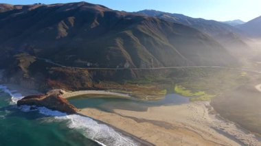 Sahne 1, Pasifik Okyanusu üzerindeki Big Sur California kayalıklarından geçiyor. Altın güneş ışığı engebeli dağları, kumlu sahili ve dramatik kıyı manzarasını ön plana çıkarıyor.