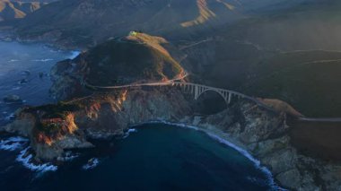 Büyük Sur sahil köprüsünün havadan görünüşü, Bixby Creek Köprüsü 'nde yıkanan altın saat ışığı ve engebeli uçurumlar, Pasifik dalgaları korunaklı körfeze çarpıyor, sahil yolunda sis sürükleniyor, süpürüyor.