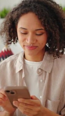 Genç siyahi bir kadın kanepede oturuyor ve evinde akıllı bir telefon kullanıyor. Rahat bir yaşam tarzı. Doğal gün ışığı, yumuşak renkler, rahat bir oturma odası ve sakin bir günlük atmosfer..