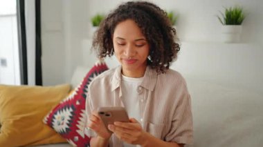 Kanepede oturan ve evinde akıllı telefon kullanan genç bir kadın, rahat bir yaşam tarzı, doğal gün ışığı, yumuşak renkler, rahat bir oturma odası ve sakin bir günlük atmosfer..