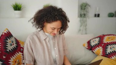 Evdeki kanepede dinlenen gülümseyen kadın, modern parlak iç mekanda huzurlu bir yaşam tarzı, doğal gün ışığı, rahat atmosfer, günlük ev kıyafetleri ve kapalı alandaki pozitif hava.