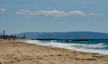 Los Angeles, California sandy ocean shore, industrial zone on the horizon, waves rolling onto the shore.