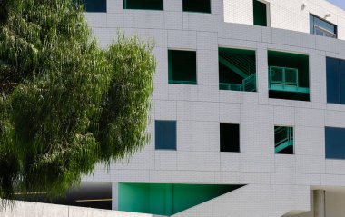Parlak şehir mimarisi altında geometrik pencereleri ve yeşil iç merdivenleri olan modern beyaz bina cephesi.