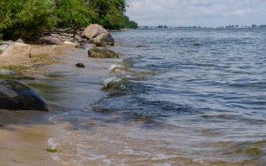 Ağaçları ve yumuşak dalgaları olan kayalık deniz kıyıları dramatik bulutlu gökyüzü altında yaz manzarası.
