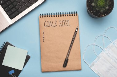 Yeni yıl hedefleri 2021. Aksiyon metni, dizüstü bilgisayar, kırtasiye malzemeleri ve açık mavi maske takan bir not defteri. Yeni normal, koronavirüs salgını. Üst görünüm, düz görünüm, kopyalama alanı