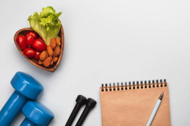 Sağlıklı yaşam tarzı ve diyet konsepti. Kalp şeklinde tabak, sebze ve fındık, spor aksesuarları, açık mavi arka planda boş not defteri. Üst görünüm, düz görünüm, kopyalama alanı