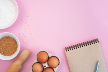 Paskalya yemeği malzemeleri ve açık pembe arka planda not defteri. Yumurta, un, merdane, şeker ve renkli konfeti. Yemek konsepti ve tatil. Üst görünüm, düz görünüm, kopyalama alanı
