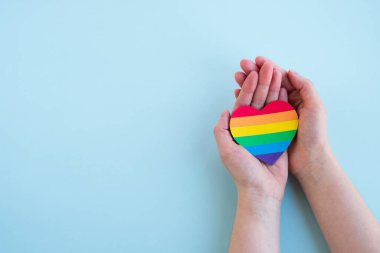 Kadınların elleri açık mavi arka planda gökkuşağının renginde kalbi tutar. LGBT konsepti, Gurur ayı. Üst görünüm, alanı kopyala