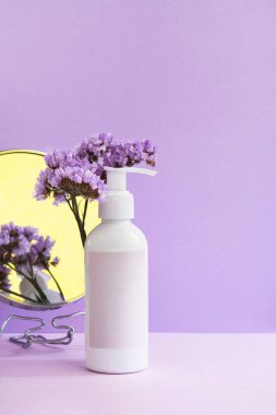 Boş kepekli kozmetik şişesi, çiçekler ve pastel mor arka planda yansıması olan ayna. Güzellik, cilt bakımı ve doğal bitkisel kozmetik. Paketleme modeli. Dikey, minimal, durgun yaşam
