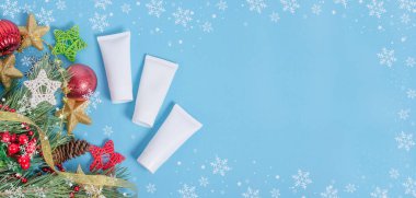 Baner özgeçmişi - Noel kozmetik satışı. Mavi arka planda Noel süslemesi olan krem tüpleri. Fotokopi alanı olan kar taneleri çerçevesi.