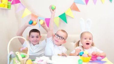 Tavşan kulaklı neşeli çocuklar Paskalya için boyalı yumurtalarla oynuyorlar. Çocuklar tatlı yer ve gülerler. İki oğlan ve küçük bir kız.