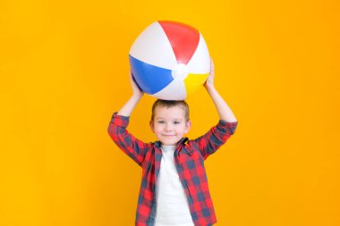 Yaz tatili konsepti, mutlu küçük çocuk portresi, gülümseyen ve elinde plaj topu tutan çocuk, şişme topla eğlenen çocuk, sarı arka planda izole edilmiş stüdyo çekimi.