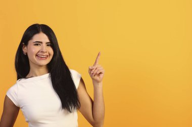 Mutlu genç bir kadın gülümsüyor ve işaret ediyor, kendine güvenen ifadesiyle boş fotokopi alanını gösteriyor, turuncu arkaplan hakkında pozitif yön ya da önemli bilgiler veriyor.