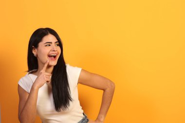 Uzun siyah saçlı, geniş bir gülümseme ile genç bir kadın ve bakışlarını başka yöne çevirerek, bir fikir düşünüyor ya da fotokopi alanı ile canlı sarı bir stüdyo arka planında sürpriz hissediyor.