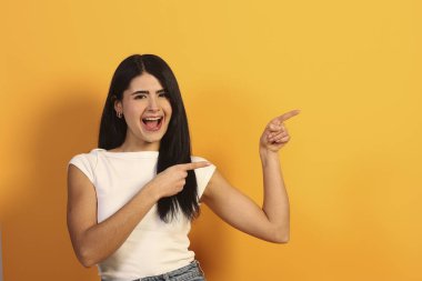 Mutlu genç bir kadın gülüyor ve coşkuyla parmaklarını sağa gösteriyor, bir fikir sunuyor veya bir ürün öneriyor, canlı sarı bir arka plana karşı duruyor.