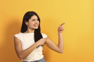 Mutlu genç bir kadın büyük bir gülümsemeyle parmaklarını kenarı işaret ediyor, boş fotokopi alanında canlı sarı bir arkaplan üzerinde bir şey gösteriyor ya da sunuyor, heyecan ve reklam veriyor.