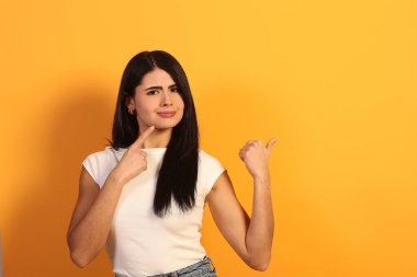 Stüdyoda sarı arka planda poz veren genç bir kadın, başparmak hareketi yaparken yüzüne işaret ederek zor bir seçim ya da düşünceli bir karar sürecini işaret ediyor.