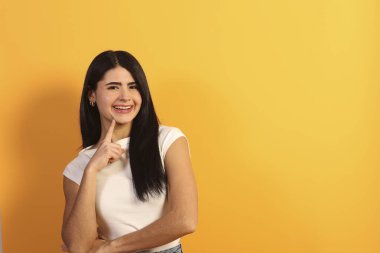 Siyah saçlı ve beyaz tişörtlü genç bir kadın düşünürken mutlu bir şekilde gülümsüyor. Fotokopi alanı olan parlak sarı bir arka plan karşısında duruyor. Fikirleri ve yaratıcı düşünceleri yansıtıyor.