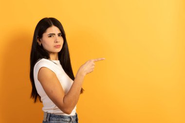 Sağı gösteren uzun siyah saçlı genç bir kadın, ciddi ve şüpheci bir yüz ifadesi sergilerken, parlak sarı arka planda geniş bir kopya alanı olan bir şey gösteriyor.