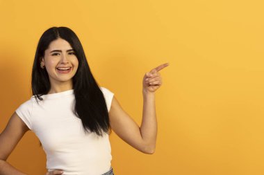 Beyaz tişörtlü, gülümseyen ve sağ elini gösteren mutlu genç bir kadın, canlı sarı arka planda geniş bir kopya alanı ile promosyon içeriği sunuyor.