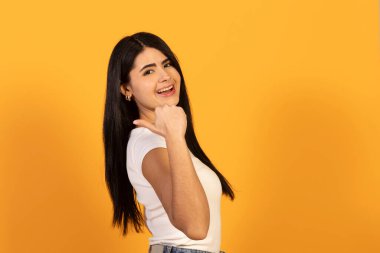 Gülümseyen, el kol hareketi yapan genç bir kadın, geriye doğru işaret ederken, parlak sarı bir arka plan üzerinde durarak, onayı, yönü, tavsiyeyi ve pozitifliği temsil eder.