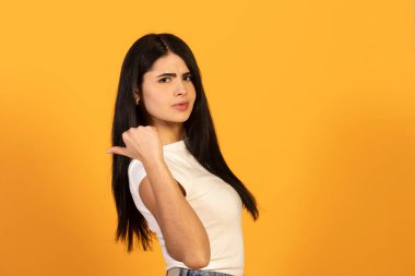 Uzun siyah saçlı, beyaz tişörtlü bir kadın, baş parmağını geriye doğru gösteriyor ve şüphe, şüphecilik ve turuncu bir arka plana karşı kafa karışıklığını ifade ediyor.