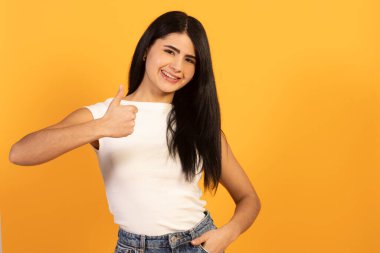 Gülümseyen genç bir kadın, onaylama, anlaşma ve memnuniyeti ifade eden, canlı sarı bir arka plana karşı özgüvenle duran,